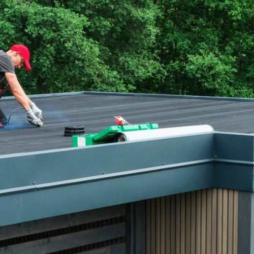 A worker in a red cap and gloves applies the best roofing materials on a flat roof of a Utah home, surrounded by trees. Tools and supplies are placed nearby.