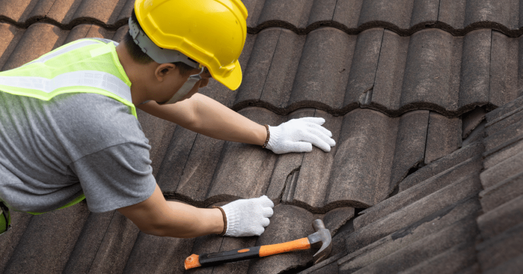 A worker in a yellow hard hat and safety vest repairs roof tiles, using gloved hands; a hammer rests nearby on the roof during an emergency roof repair.