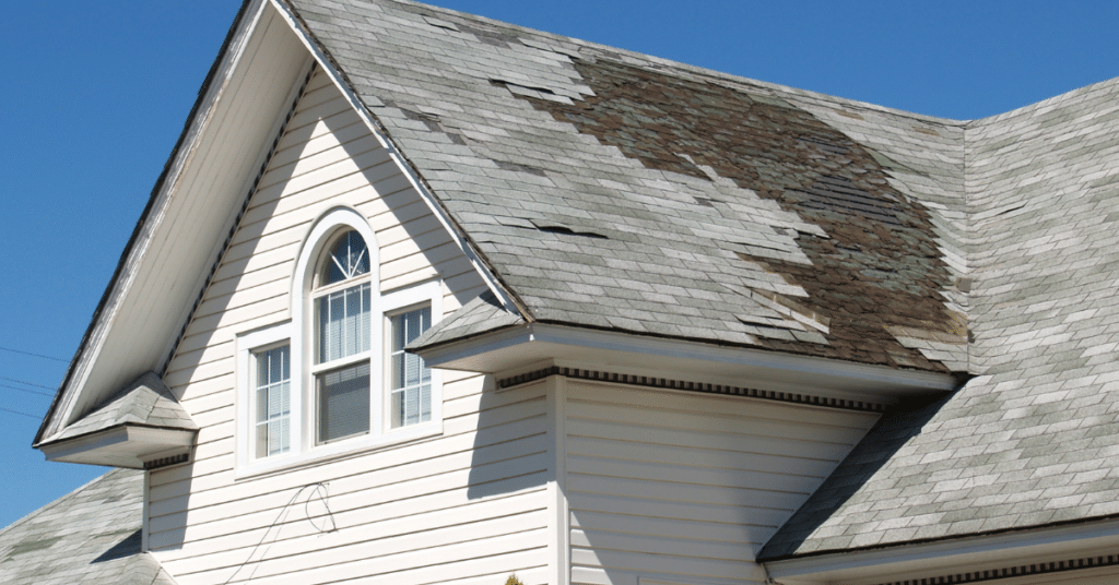 A house with white siding and a steep roof shows extensive shingle damage and missing shingles on one side, highlighting the urgent need for emergency roof repair under a clear blue sky.