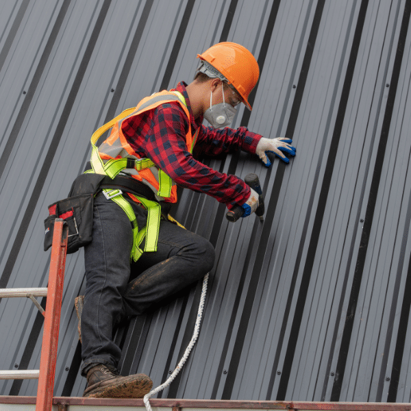 A construction worker wearing safety gear installs metal panels on a sloped roof using a power tool, showcasing skilled labor often included in the average metal roof price for homes in Utah.