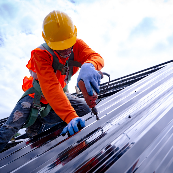 A construction worker wearing safety gear uses a power drill to install metal roofing panels on a building under a cloudy sky.