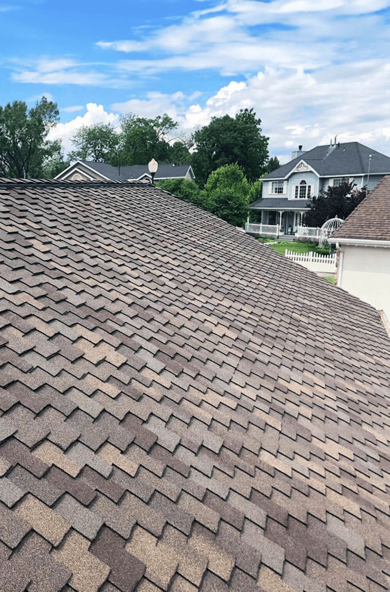 View of residential rooftops with brown asphalt shingles against a backdrop of trees and neighboring houses under a partly cloudy sky.